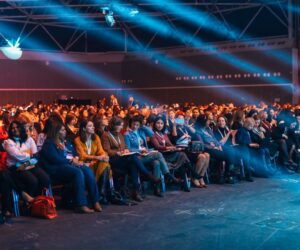 Vibrant image of a diverse audience seated in a large auditorium, highlighted by dynamic stage lighting.