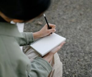 Person writes in a blank spiral notebook with a pencil outdoors.