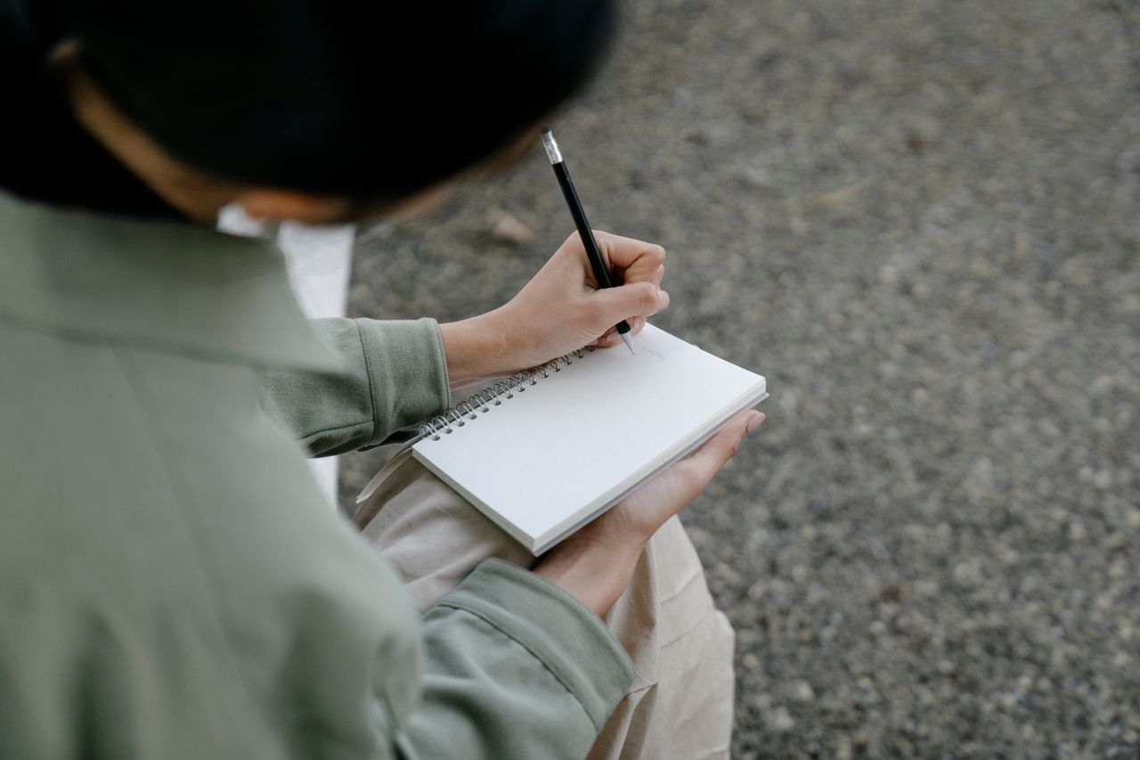 Person writes in a blank spiral notebook with a pencil outdoors.